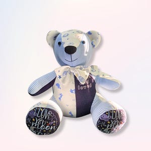 May include: A patchwork teddy bear in blue, white, and purple. The bear has a bow tie and the words "I love you to the moon" on its feet. A soft toy, the bear is made from various patterned fabrics.