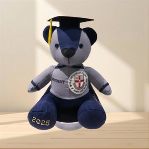 May include: A plush teddy bear wearing a black graduation cap with a yellow tassel. The bear is navy blue and grey, featuring the embroidered text "STUDENT" and "2025" along with a circular patch for the University of Chester Faculty of Health and Social Care.