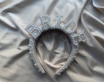 May include: A white headband with the word "BRIDE" spelled out in pearl beads. The headband is on a white fabric background.