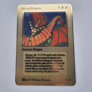 May include: A gold-colored Shivan Dragon trading card featuring a red dragon illustration against a dark blue starry background. The card text includes Summon Dragon, Flying, and a flavor text description about the dragon's cruel nature. Illus. Melissa Benson, 5/5.