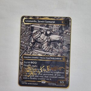 May include: A rectangular trading card titled Leonardo, Sewer Samurai featuring a black and white illustration of a turtle character with swords. The card has a gold border, black text boxes, and a gold signature across the lower portion of the card.