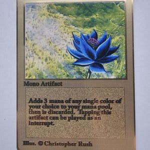 May include: A gold-colored trading card titled Black Lotus with a cost of 0. The card features a blue lotus flower illustration, the text Mono Artifact, and game rules stating it adds 3 mana of any single color. It is credited to Christopher Rush.