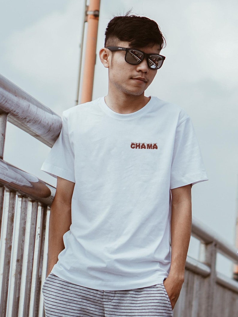 Op de afbeelding: Een man draagt een wit T-shirt met het woord "CHAMA" erop gedrukt in het rood. Hij draagt een zwarte zonnebril en heeft kort zwart haar. Hij staat op een metalen loopbrug.