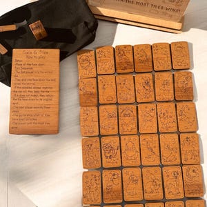 May include: A wooden game set with 24 tiles featuring different animals. Each tile has a star design and the game instructions are printed on a separate wooden board. The game is called "Tails & Tiles" and the instructions say to "Roll the Die!" and "Player with the most tiles wins!"