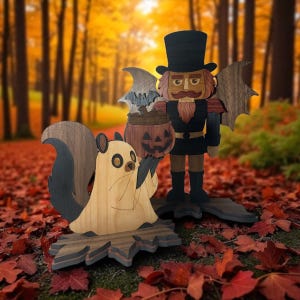 May include: Wooden Halloween decorations featuring a squirrel in a ghost costume and a nutcracker with bat wings. The squirrel is light brown with a black tail, and the nutcracker holds a pumpkin. The background is an autumn forest.