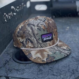 May include: A baseball cap featuring a brown, green, and tan camouflage leaf pattern. The cap has a white cord across the base of the crown and a rectangular black patch on the front with a purple mountain graphic and the text pownal in white.