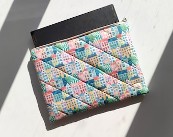 Op de afbeelding: Een gewatteerde laptoptas met een kleurrijk dorpspatroon. De tas is open en onthult een zwarte laptop. Het patroon toont rijen huizen in verschillende kleuren. Met ritssluiting.