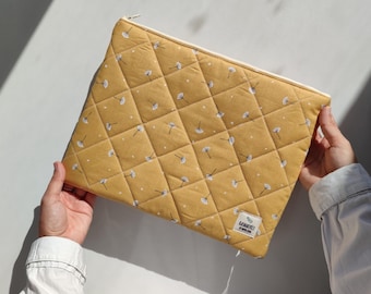 Op de afbeelding: Mosterdgele gewatteerde etui met een wit bloemmotief en een ruitpatroon. De etui heeft een ritssluiting en een klein label. Afmetingen in centimeters. De etui wordt door een persoon vastgehouden.