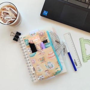 Puede incluir: Un cuaderno de espiral con un bolsillo temático de té, lápices y un bolígrafo. También se ve un vaso de café helado y un ordenador portátil. Un transportador, una regla y otros artículos de papelería están dispuestos sobre una superficie blanca.