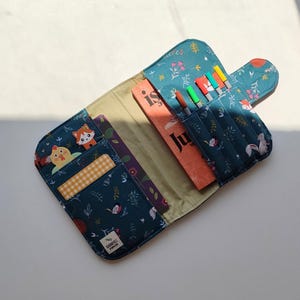 May include: Open teal fabric pencil case with a whimsical forest animal print. The case is open, revealing a book, colored markers, and pockets. The case has a button closure and is lined with a light green fabric.