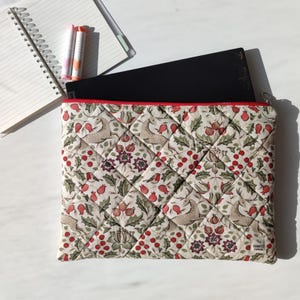 Puede incluir: Funda acolchada para portátil con ribete rojo, con un estampado repetido de pájaros, bayas y follaje en tonos rojos, verdes y beige. Un portátil negro es parcialmente visible en el interior. Un cuaderno y bolígrafos están en el fondo.