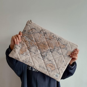 Könnte beinhalten: Eine gesteppte Clutch in Beige und Braun mit Weltkarte und Reißverschluss. Die Kontinente und Ozeane sind mit schwarzer Schrift beschriftet. Die Clutch wird vor einer Person gehalten.