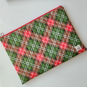 May include: A quilted plaid zippered pouch in red, green, white, and brown. The pouch features a diamond pattern and a red zipper. A small white tag with text is sewn on the bottom right corner. The pouch is likely used for storing items.