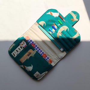 May include: An open teal pencil case with a dachshund pattern. The case has multiple compartments, including slots for pens and pencils, and pockets for small items. A cat bookmark is visible in one of the pockets.