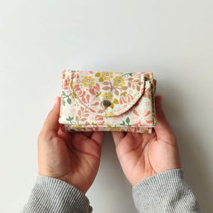 May include: A small, floral-patterned wallet with a snap closure. The wallet features a design of pink, yellow, and green flowers on a cream background. The wallet is held open, showcasing its interior. The overall aesthetic is delicate and feminine.