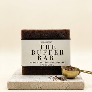 May include: A dark brown bar of soap labeled "THE BUFFER BAR" with "PUMICE • WALNUT SHELL POWDER" and a net weight of 4.5 oz / 130 g. A small gold scoop and a pile of brown powder sit on a stone block.