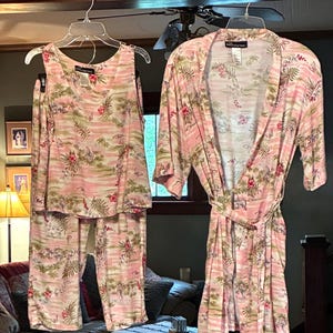 Puede incluir: Un conjunto de pijama rosa y una bata a juego con estampado floral tropical. El conjunto incluye una camiseta sin mangas y pantalones. La bata tiene un cinturón. Todos los artículos están colgados en perchas transparentes.