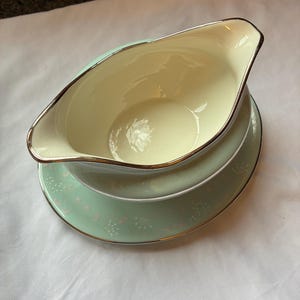 May include: A vintage cream-colored gravy boat with a gold rim, resting on a matching plate. The plate is light green with a delicate white floral pattern and a gold rim. The set is likely porcelain.