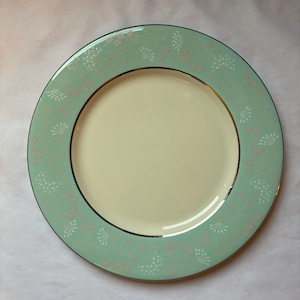 May include: A round, ceramic plate with a pale yellow center and a mint green border. The border features a delicate floral pattern in white and pink, with a thin gold and black trim. This decorative plate is suitable for serving food.
