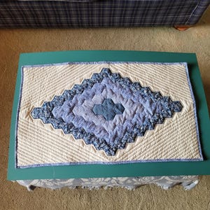 May include: A quilted table runner with a central diamond pattern in shades of blue and white. The diamond is surrounded by a cream-colored background with diagonal stitching. The runner is edged with a blue border.