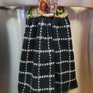 May include: Black and white checkered kitchen towel with a floral print fabric loop. The loop has a button and the text "DAISY" and "GERBERA" is visible.