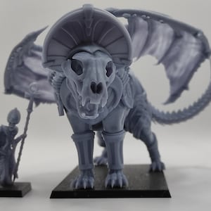 Puede incluir: Una miniatura gris impresa en 3D de una criatura esquelética similar a un lobo con alas y un tocado. La criatura está de pie sobre una base negra. Una miniatura más pequeña de color gris impresa en 3D de una figura esquelética con un bastón está de pie sobre una base negra en primer plano.
