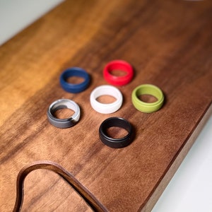 May include: Seven silicone rings in different colours, including black, white, grey, red, blue, and green. The rings are arranged on a wooden surface.