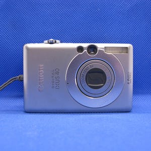 May include: Silver Canon Digital IXUS 40 camera with a 3x zoom lens. The camera has a built-in flash and a wrist strap. The camera is on a blue background. The camera has the text "Canon DIGITAL IXUS 40" printed on the front.