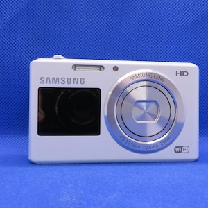 May include: A white Samsung digital camera with a silver lens and a black viewfinder. The camera has a silver button on the front and the text "SAMSUNG LENS" and "HD" printed on the top. The camera also has the text "4.5-22.5mm 1:2.5-6.3 28mm" and "Wi-Fi" printed on the front.