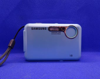 May include: A light blue Samsung digital camera with a black strap. The camera has a silver lens with the text "SAMSUNG LENS 3.5-4.5/6.3-18.9mm" printed on it. The camera also has the text "i8" printed on the bottom right corner.