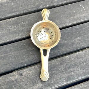 Puede incluir: Un colador de té de metal dorado con mango. El colador tiene un cuenco redondo perforado y un mango decorativo.