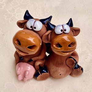 Può includere: Una statuetta in ceramica raffigurante due mucche marroni con grandi occhi espressivi e corna nere. Una mucca ha una mammella rosa. Le mucche sono sedute vicine, creando un pezzo decorativo stravagante e affascinante.