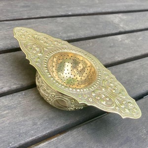 Könnte beinhalten: Ein goldfarbener Teesieb aus Metall mit einem verzierten Design. Das Sieb hat eine runde, perforierte Mitte und einen dekorativen, gewellten Rand.