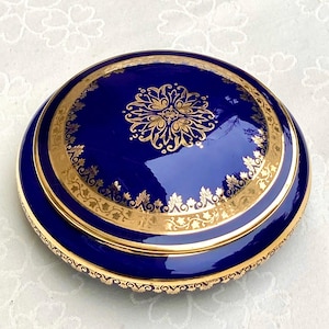 Può includere: Un portagioie rotondo blu cobalto con un motivo floreale dorato sul coperchio. La scatola ha un bordo dorato e un bordo decorativo dorato. La scatola è probabilmente in porcellana o ceramica.