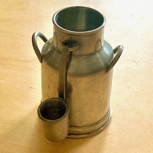 May include: A small, antique-style metal milk can with a matching small cup attached. The can has two arched handles and a spout. The metal has a dull, aged appearance. The can is sitting on a light-colored wooden surface.