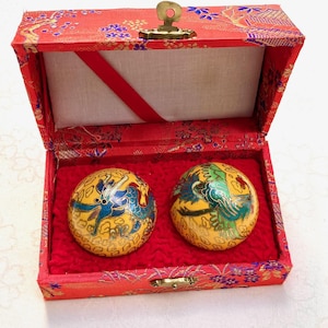 Può includere: Una scatola rossa a fantasia con chiusura dorata, contenente due ornamenti rotondi e gialli. Ogni ornamento presenta un colorato disegno di drago o uccello. La scatola ha un interno rosso e una fodera color crema.
