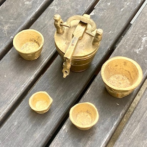 Op de afbeelding: Een antieke messing vijzel en stamper set met vier kleine, lichtgekleurde kommen. De vijzel heeft een scharnierend deksel en een lange handgreep. De set wordt gepresenteerd op een verweerd houten oppervlak.