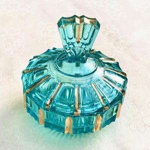 Op de afbeelding: Een turquoise glazen sieradendoosje met deksel. Het heeft een geometrisch ontwerp met gouden accenten. De deksel heeft een facetgeslepen, waaiervormige handgreep. De diameter is ongeveer 10 cm.