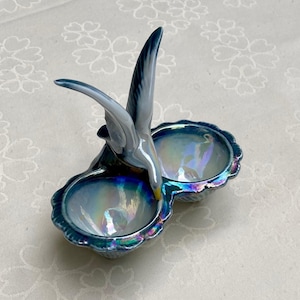 Può includere: Un piatto doppio in ceramica blu iridescente con una statuetta di uccello in cima. Il piatto ha la forma di due conchiglie.