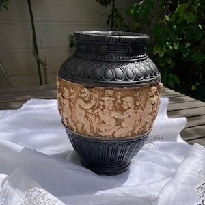 Könnte beinhalten: Eine schwarze Keramikvase mit einem Reliefmuster von spielenden Cherubim. Die Vase steht auf einer weißen Leinen-Tischdecke mit Spitzenbesatz.