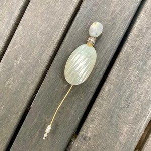 Könnte beinhalten: Ein weißer, perlmuttartiger Flaschenverschluss mit einer goldenen Nadel. Der Flaschenverschluss hat ein strukturiertes, geripptes Design.