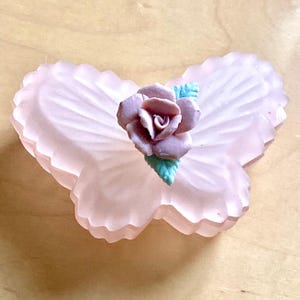 Könnte beinhalten: Eine hellrosa, schmetterlingsförmige Schale mit mattierter Oberfläche. Eine detaillierte, malvenfarbene Rose mit türkisfarbenen Blättern befindet sich in der Mitte der Schale. Die Schale ist wahrscheinlich aus Glas oder einem ähnlichen Material.