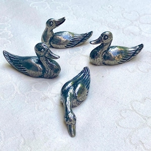 Peut inclure: Quatre figurines de canards argentés. Les canards sont dans différentes poses, avec des textures de plumes et des becs détaillés. Les figurines sont probablement des objets décoratifs, peut-être pour une table ou une exposition.