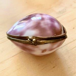 Può includere: Un piccolo portagioie di forma ovale realizzato in porcellana lucida e marmorizzata con sfumature vorticose di bianco e viola intenso. Il contenitore presenta un bordo in ottone incernierato con un semplice meccanismo di chiusura a scatto.