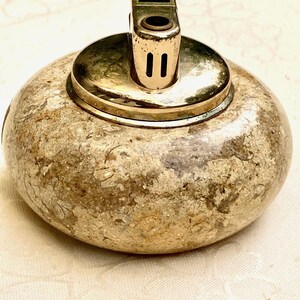 May include: A vintage lighter with a gold-coloured metal top and a textured, stone-like base. The top of the lighter has the word "ATOMIC" printed on it. The lighter is round and sits on a white surface.