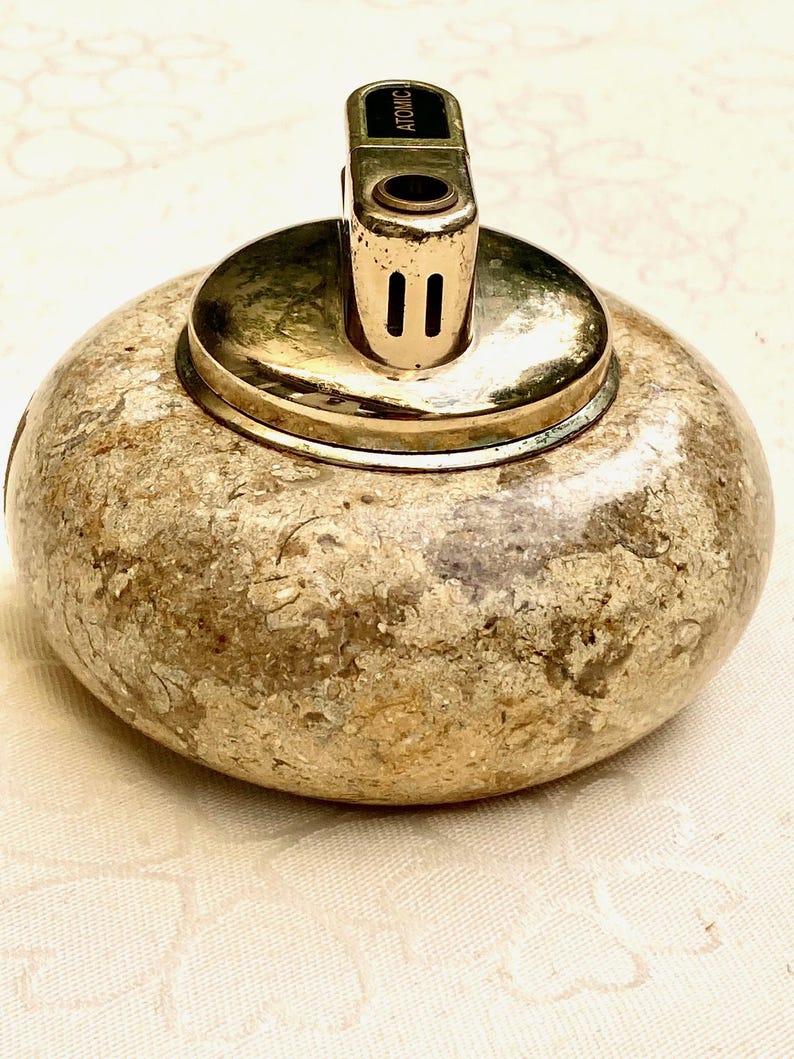 May include: A vintage lighter with a gold-coloured metal top and a textured, stone-like base. The top of the lighter has the word "ATOMIC" printed on it. The lighter is round and sits on a white surface.