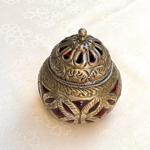 Peut inclure: Un récipient en métal argenté et rouge avec un design orné et un couvercle en dôme. Le récipient a un insert en verre rouge et des motifs floraux complexes.