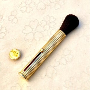 Può includere: Un pennello per trucco dorato con una testina marrone scuro e un cappuccio abbinato. L'impugnatura del pennello presenta scanalature verticali e un piccolo gioiello decorativo. Il cappuccio è rotondo e ha una superficie giallastra.