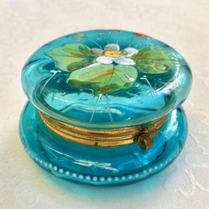 Könnte beinhalten: Eine türkisfarbene Glas-Schmuckschatulle mit einem Blumendesign auf dem Deckel. Der Deckel zeigt eine weiße Blume mit grünen Blättern. Die Schatulle hat ein goldfarbenes Metallband und einen Verschluss. Der Boden der Schatulle hat ein weißes Punktmuster.