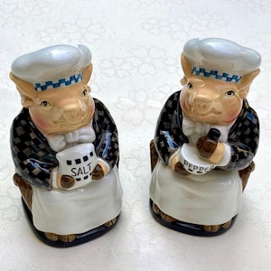 Peut inclure: Deux salières et poivrières en céramique en forme de cochons. Chaque cochon porte un chapeau de chef avec une bande à carreaux bleus et blancs, une veste à carreaux noirs et blancs et un tablier blanc. L'un tient un sac avec "SEL", l'autre un poivrier.